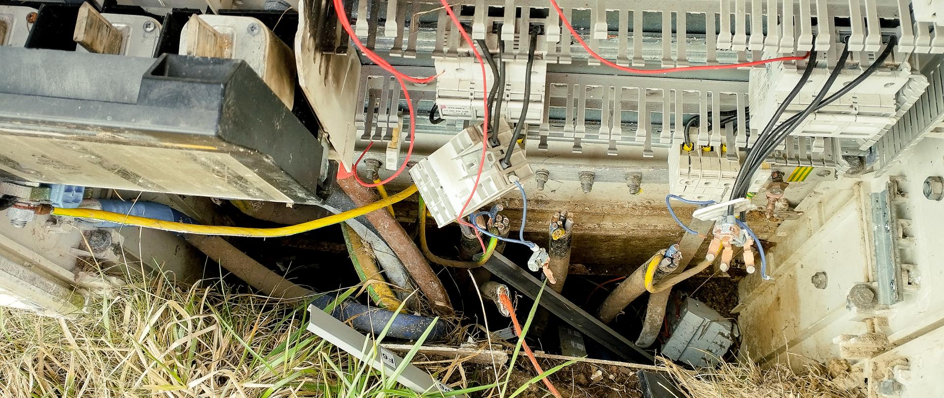 Na fotografii widzimy otwartą skrzynkę oświetleniową, która została uszkodzona w wyniku kradzieży przewodów. Wewnątrz znajdują się porozrzucane i pocięte kable różnych kolorów, wskazujące na celową ingerencję. Stan skrzynki sugeruje, że złodzieje usunęli znaczną część miedzianych przewodów, prawdopodobnie w celu ich sprzedaży. Na zewnątrz skrzynki widać trawę i ziemię, co sugeruje, że skrzynka znajduje się na otwartej przestrzeni. Całość pozostawia wrażenie nieuporządkowania i zaniedbania, co podkreśla skutki aktu wandalizmu.
