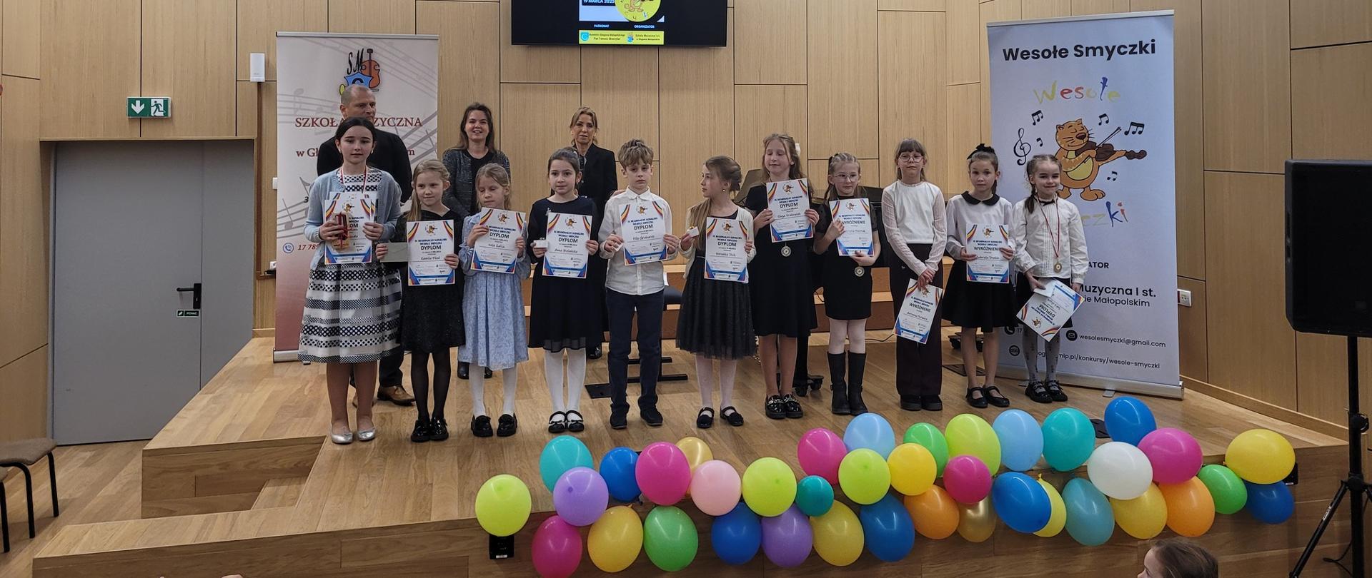 Na scenie w nowoczesnej sali koncertowej stoi grupa dzieci ubranych w eleganckie stroje, trzymających dyplomy i nagrody. W tle widoczne są banery konkursowe, w tym jeden z napisem „Wesołe Smyczki” oraz grafiką misia grającego na skrzypcach. Za dziećmi stoją nauczycielki, uśmiechające się dumnie. Przed sceną znajdują się kolorowe balony, a w dolnym rogu zdjęcia ktoś robi fotografię telefonem. W sali panuje radosna atmosfera, a dzieci wyglądają na szczęśliwe i dumne ze swoich osiągnięć.