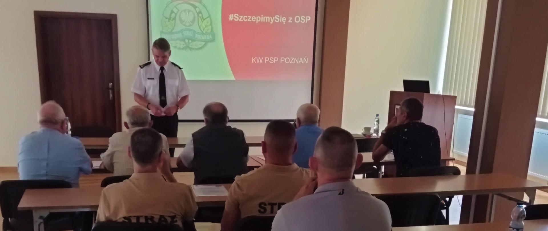Zdjęcie przedstawia strażaków podczas spotkanie w świetlicy komendy, stojących lub siedzących na krzesłach.
W tle prezentacja multimedialna na ekranie ściennym. 