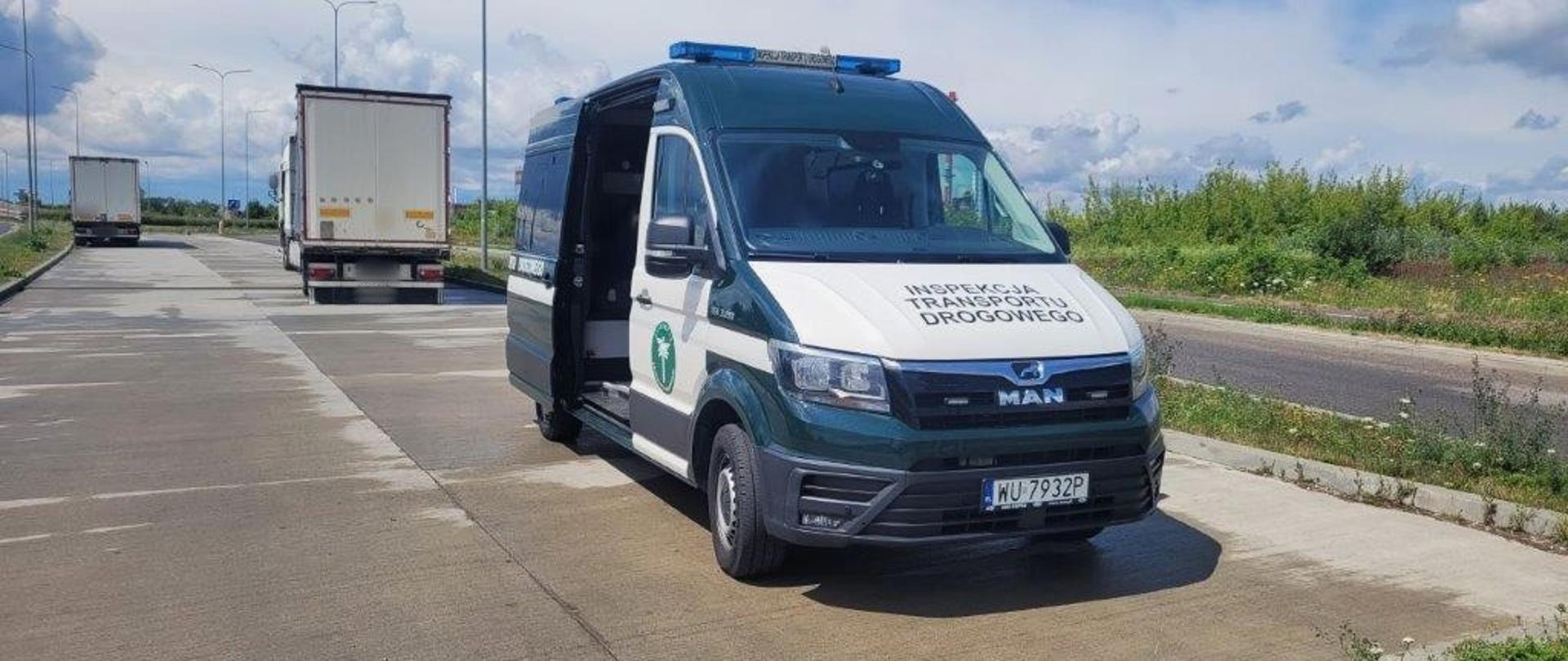 Na pierwszym planie (od prawej): przód i prawy bok oznakowanego furgonu mazowieckiej Inspekcji Transportu Drogowego. Za radiowozem Inspekcji Transportu Drogowego stoi ciągnik siodłowy z podpiętą naczepą. Wszystkie pojazdy znajdują się w wyznaczonej zatoce kontrolnej przy drodze krajowej numer 9 w Radomiu. W tle: drzewa, krzewy i lokalna droga dojazdowa.