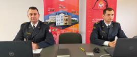 Na zdjęciu widać Komendanta Powiatowego Państwowej Straży Pożarnej w Gnieźnie siedzącego przed laptopem podczas narady rocznej. Obok niego siedzi jego zastępca. W tle stoją banery naszej komendy