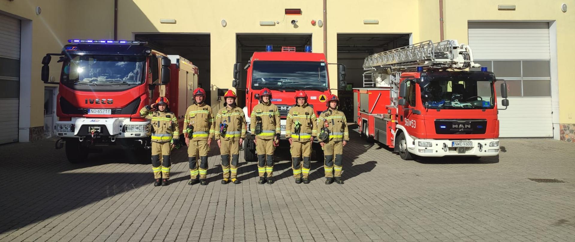 Strażacy stoją przed budynkiem KP PSP i oddają honor poległym. Za nimi stoją 3 wozy bojowe. 
