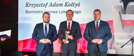 Minister Czarnek i dwóch mężczyzn w garniturach stoją, jeden z mężczyzn trzyma małą statuetkę, za nimi ścianka z napisem Krzysztof Adam Kołtyś Burmistrz Janowa Lubelskiego.