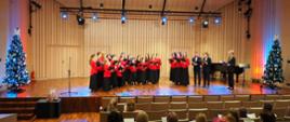 Chór młodzieżowy stoi na scenie między udekorowanymi świątecznie choinkami w sali koncertowej.