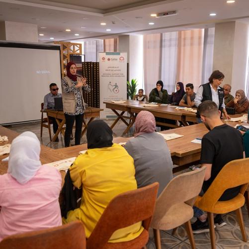 Grupa ludzi, kobiet i mężczyzn, siedzi wokół ustawionego w podkowę stołu, stojąca kobieta w chuście prowadzi spotkanie