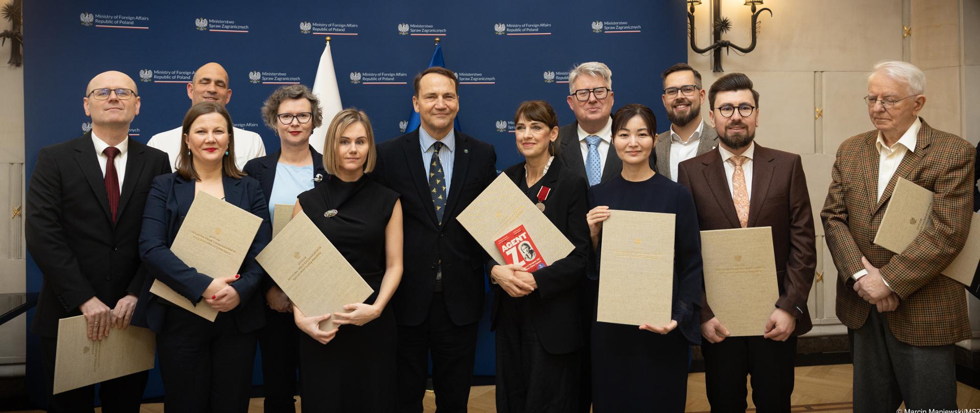 Przed ścianką prasową MSZ stoi grupa kobiet i mężczyzn trzymających dyplomy w związku z udziałem w konkursie na najlepszą książkę historyczną. 