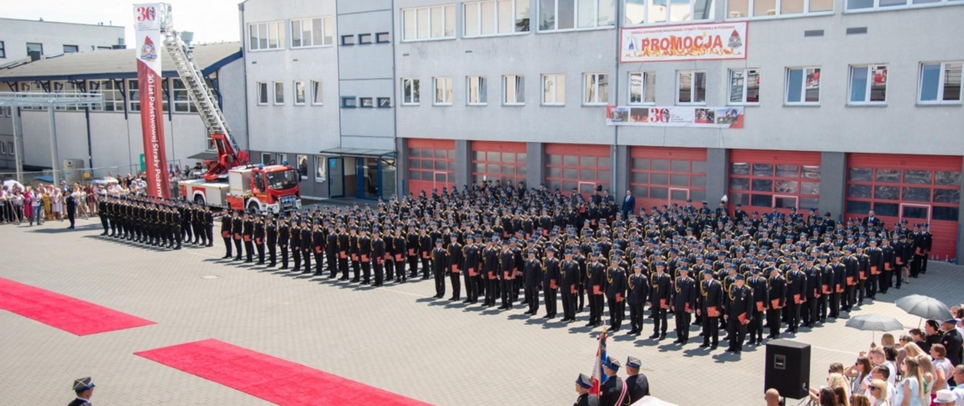 Zdjęcie przedstawia plac wewnętrzny Szkoły Aspirantów Państwowej Straży Pożarnej w Poznaniu podczas uroczystego apelu w związku z promocja aspirancką. Na zdjęciu znajdują się pododziały PSP, sztandary oraz publiczność.