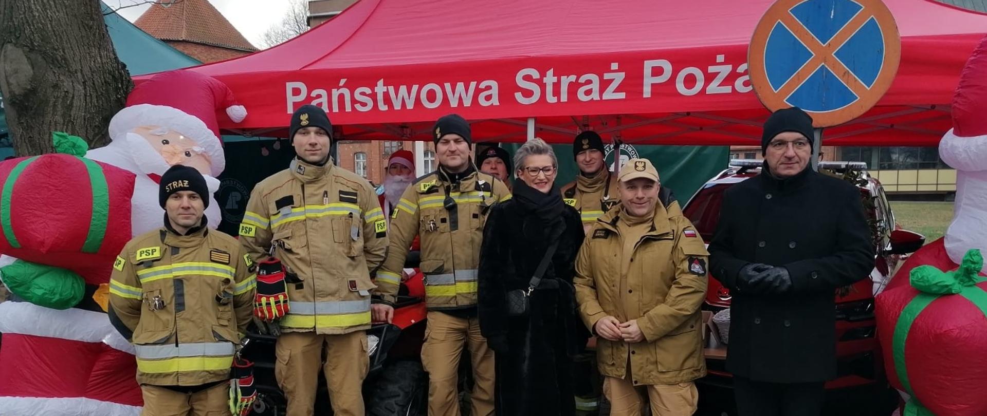 Strażacy stoją pod czerwonym namiotem z prawej strony burmistrz Braniewa. Trwa jarmark Mikołajkowy.