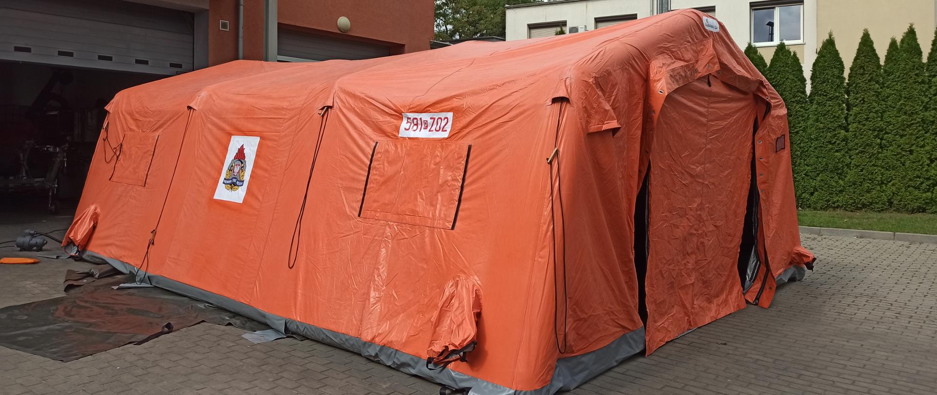 pomarańczowy namiot pneumatyczny stoi rozstawiony na kostce brukowej za nom budynek z otwarta bramą garażowa zanim zielone drzewo oraz inny budynek