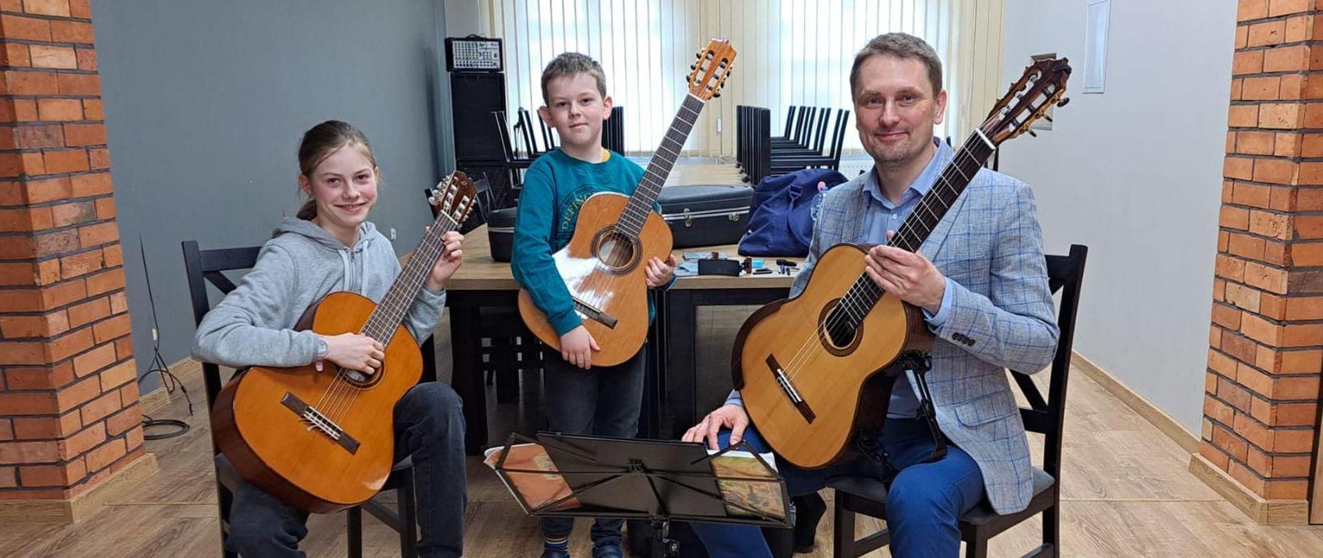 Dwoje uczniów i nauczyciel siedzą trzymając gitary. Przed nimi pulpit z nutami.
