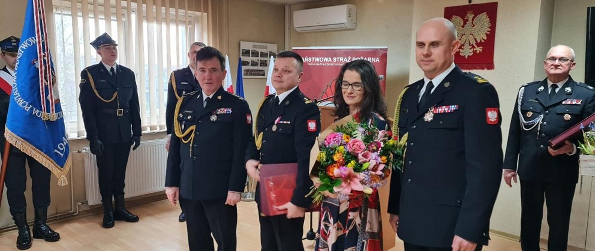 Na fotografii stoi zastępca podkarpackiego komendanta wojewódzkiego Państwowej Straży Pożarnej, st.bryg. Andrzej Marczenia, odchodzący ze służby zastępca komendanta powiatowego Państwowej Straży Pożarnej, bryg. Marek Gawlak, małżonka Pani Ewa Gawlak oraz komendant powiatowy Państwowej Straży Pożarnej, bryg. Łukasz Kapinos. Strażacy ubrani w mundury galowe koloru granatowego. W tle zdjęcia widać godło państwowe i krzyż. Po lewej stronie zdjęcia widać sztandar komendy powiatowej Państwowej Straży Pożarnej w Mielcu.