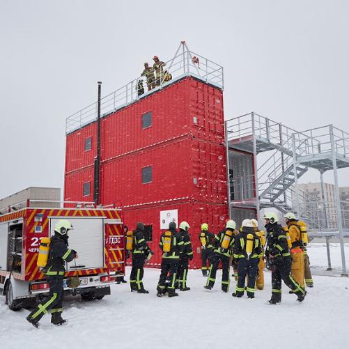 Obok trenażera stoi wóz strażacki oraz grupa strażaków i ratowników w kombinezonach i hełmach; na szczycie trenażera stoi trzech mężczyzn