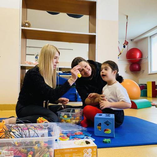 Dwie kobiety prowadzą zajęcia rehabilitacyjne z jedną dziewczynką. Wokół sporo zabawek i urządzeń rehabilitacyjnych.