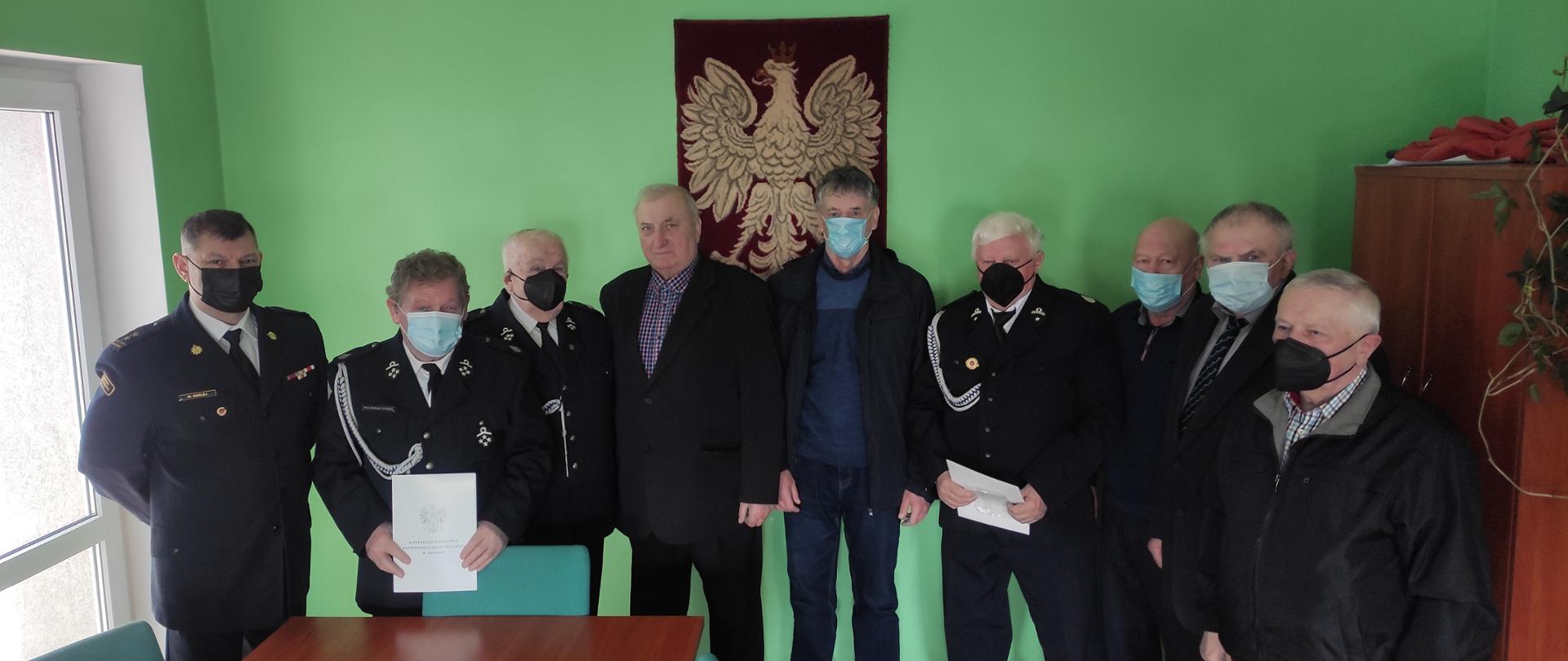 Obraz przedstawia Komendanta Powiatowego wraz ze strażakami OSP, którym przyznano świadczenie ratownicze. Osoby znajdują się w sali narad.