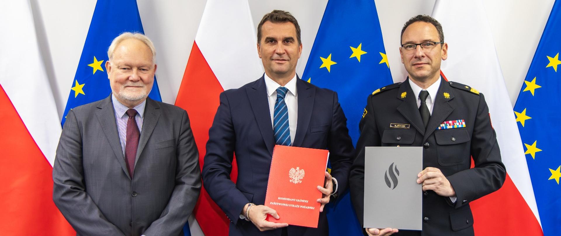 Trzech mężczyzn pozuje do zdjęcia na tle flag Polski i Unii Europejskiej. Pośrodku stoi mężczyzna w garniturze z niebieskim krawatem, trzymający czerwony segregator z godłem Polski. Po jego lewej stronie znajduje się mężczyzna w szarym garniturze z bordowym krawatem, a po prawej funkcjonariusz Państwowej Straży Pożarnej w mundurze galowym, trzymający teczkę z logo PSP.