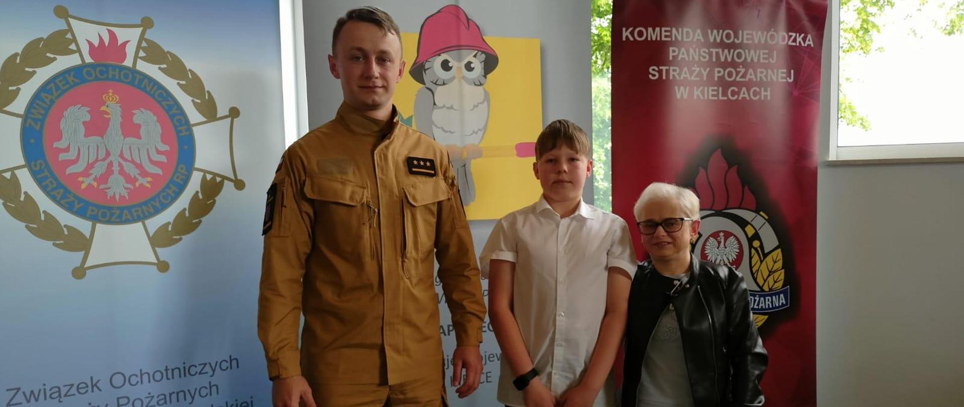 Na pierwszym planie widoczny uczestnik Ogólnopolskiego Turnieju Wiedzy Pożarniczej, po jego lewej stronie znajduje się funkcjonariusz PSP, po jego prawej stronie opiekun ze szkoły. Za nimi widoczne banery Związku Ochotniczych Straży Pożarnych RP, Ogólnopolskiego Turnieju Wiedzy Pożarniczej oraz Komendy Wojewódzkiej PSP w Kielcach.