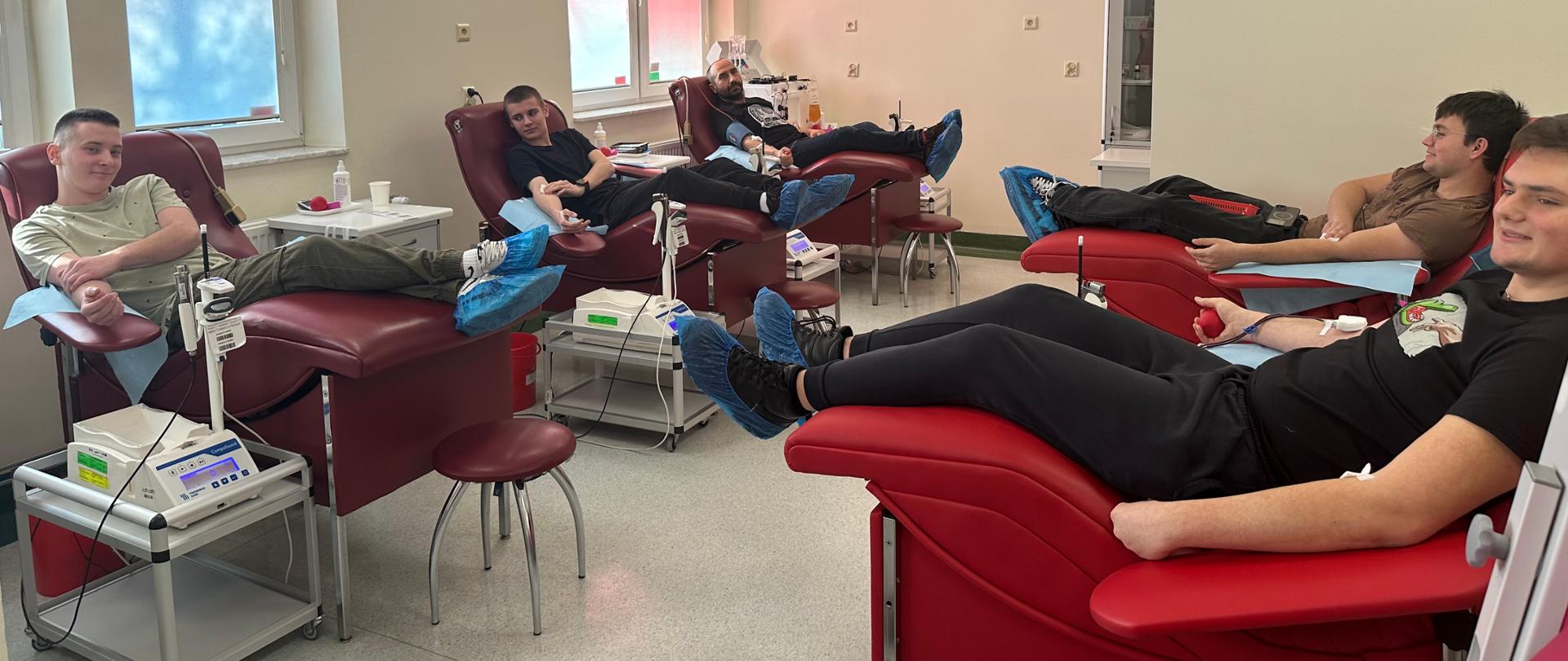 Kilka osób leży na fotelach w sali krwiodawstwa podczas oddawania krwi; widoczna aparatura medyczna
