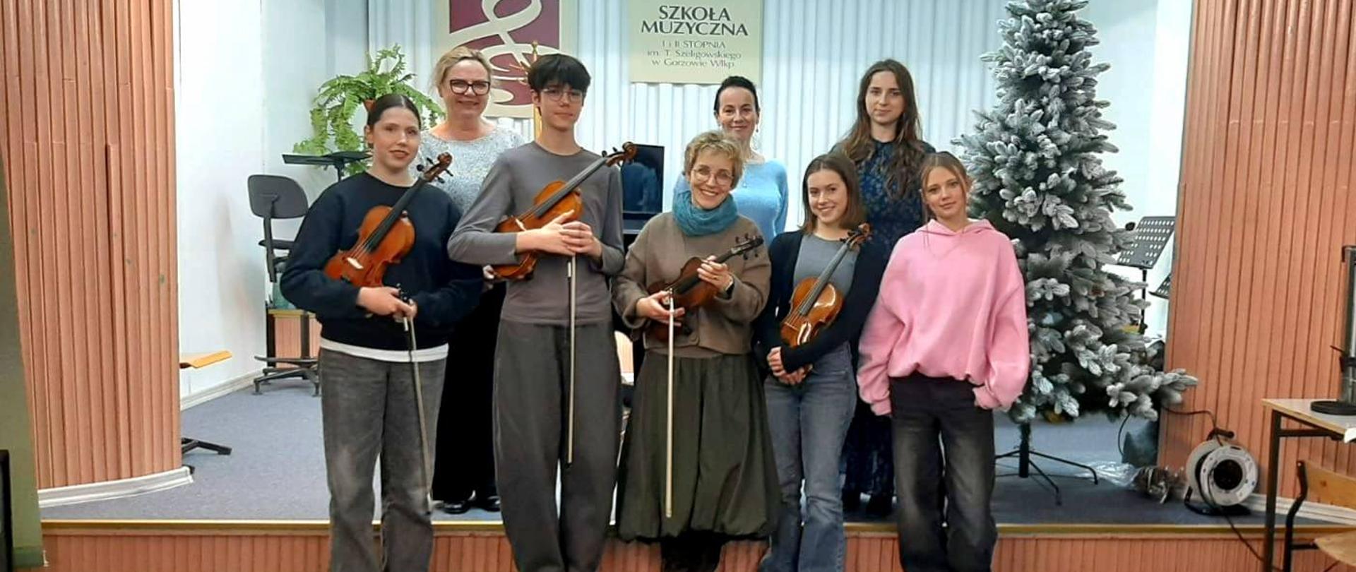 Uczniowie, nauczyciele i prowadząca warsztaty stoją na scenie ze skrzypcami