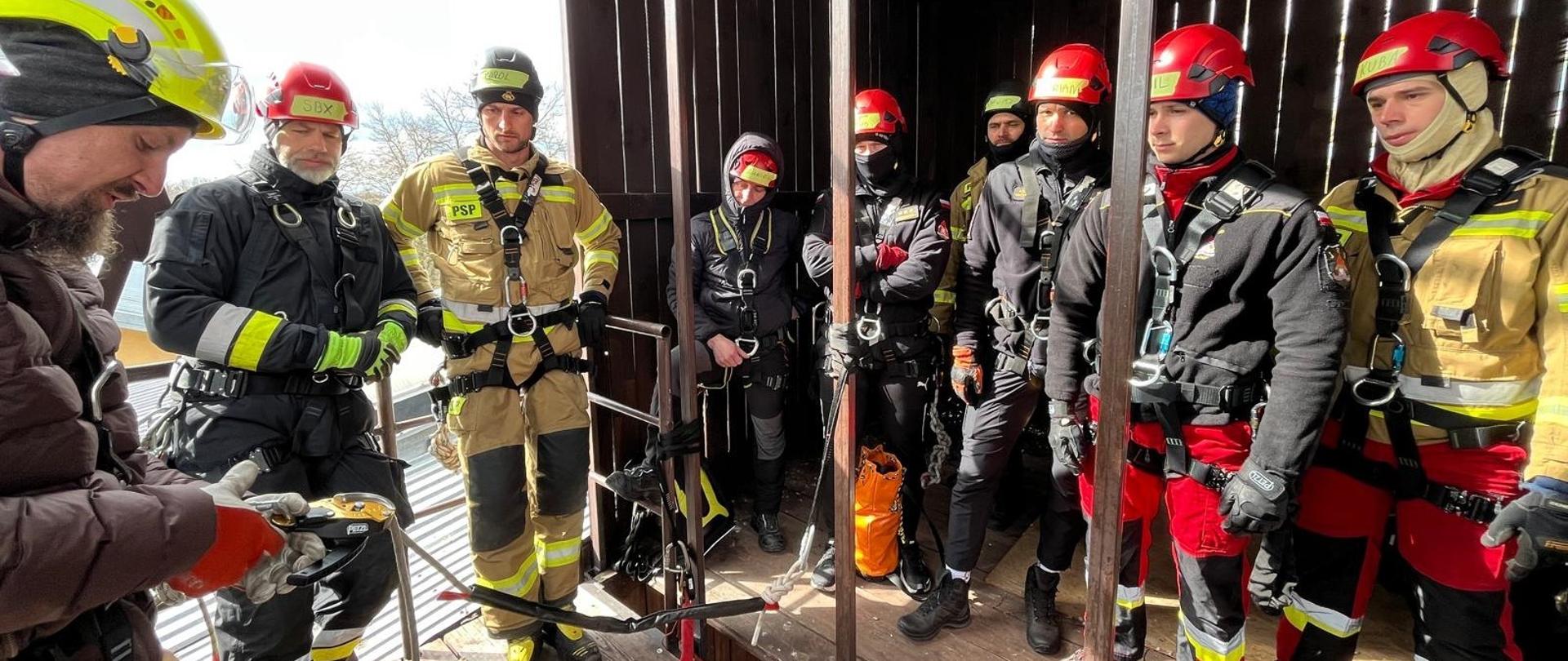 Na zdjęciu znajduje się grupa strażaków w pełnym umundurowaniu ochronnym, wyposażona w sprzęt do ratownictwa wysokościowego. Wszyscy mają założone uprzęże asekuracyjne i kaski ochronne w jaskrawych kolorach – czerwonych, żółtych i czarnych. Stoją na drewnianej platformie treningowej, osłoniętej ciemnymi drewnianymi ścianami i metalowym zadaszeniem. Po lewej stronie kadru jeden z instruktorów, ubrany w ciemną kurtkę i kask w kolorze neonowej żółci, prezentuje działanie sprzętu linowego, trzymając w dłoniach karabinki i przyrządy zjazdowe. Pozostali uczestnicy uważnie słuchają i obserwują. W tle widoczne są liny asekuracyjne oraz zwoje czerwonej liny ratowniczej leżące na ziemi. Atmosfera na zdjęciu jest skupiona i profesjonalna – to moment nauki i przygotowania do ćwiczeń praktycznych na wysokości.