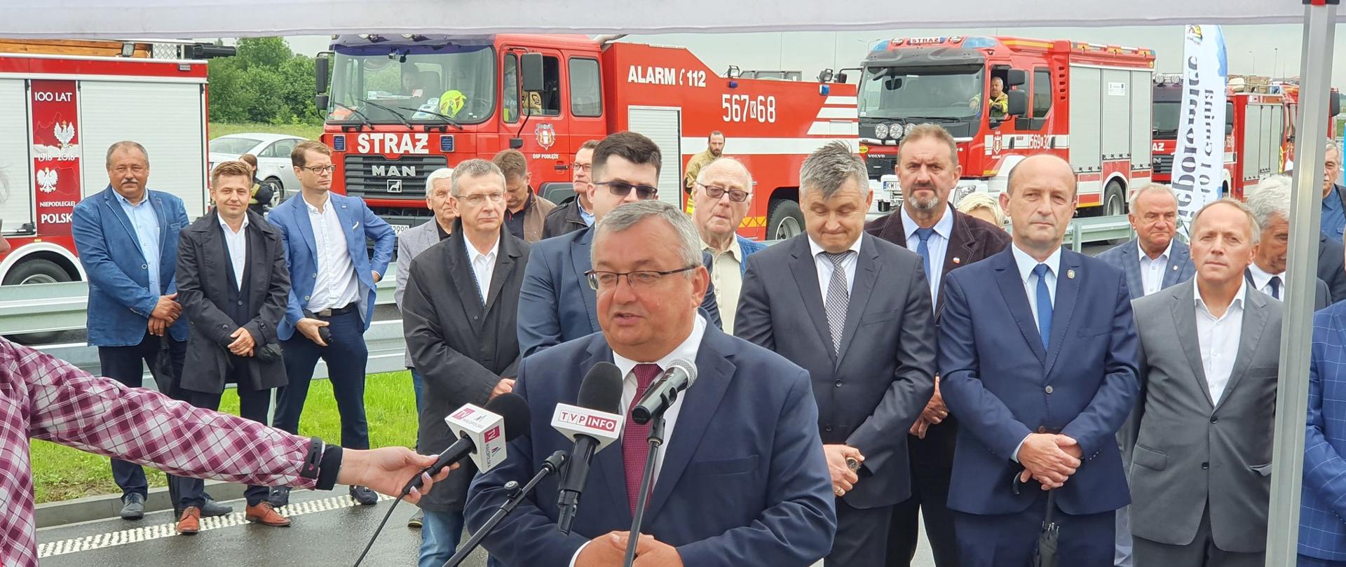 Na zdjęciu widać grupę ludzi stojących pod namiotem, w ich tle widać kilka samochodów straży pożarnej. Jeden z mężczyzn stoi przy mikrofonie. a