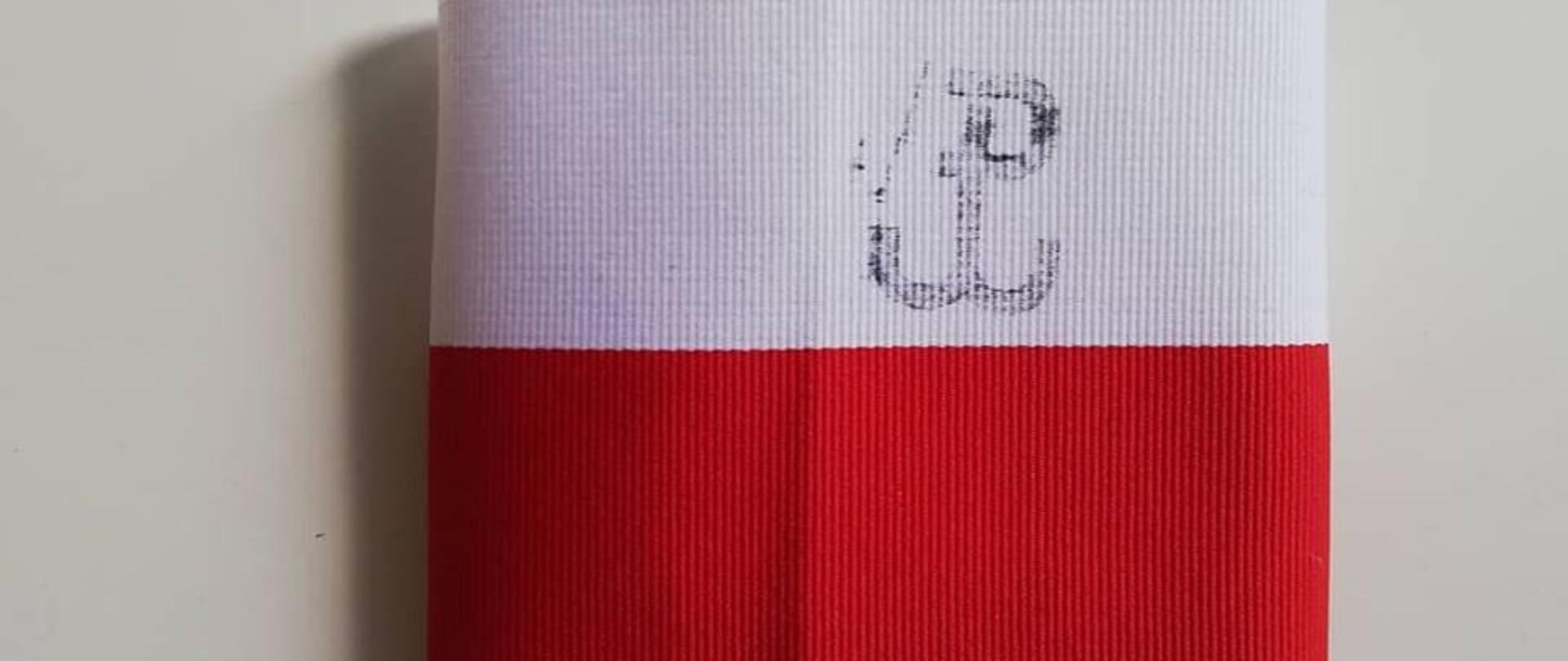 Na jasnym tle biało-czerwona opaska z kotwicą