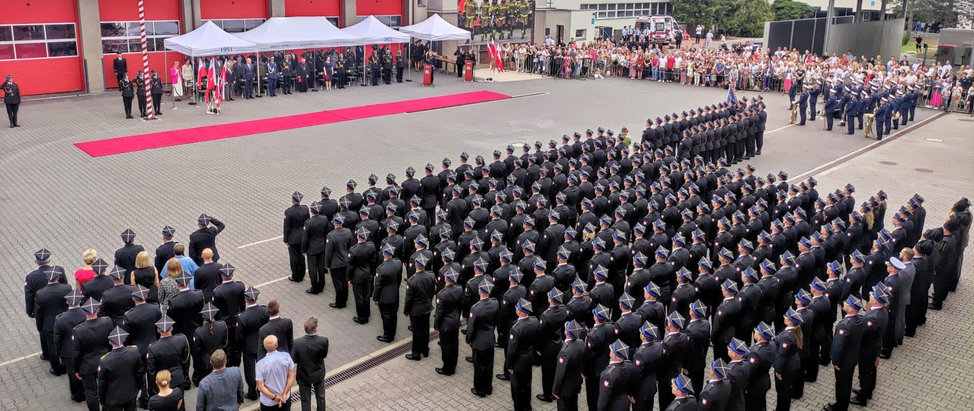 Zdjęcie przedstawia plac wewnętrzny Szkoły Aspirantów Państwowej Straży Pożarnej w Poznaniu podczas uroczystego apelu w związku z promocja aspirancką. Na zdjęciu znajdują się pododziały PSP, sztandary oraz publiczność.