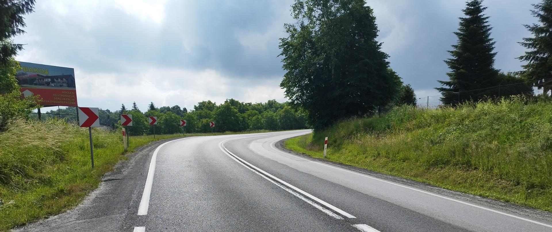 Zdjęcie zrobione w okresie letnim. Przedstawia drogę jednopasmową, po jednym pasie w każdym kierunku. Droga skręca tworząc ostry zakręt w prawo. Jest on odpowiednio oznakowany. Po lewej stronie zdjęcia jest baner reklamowy. 