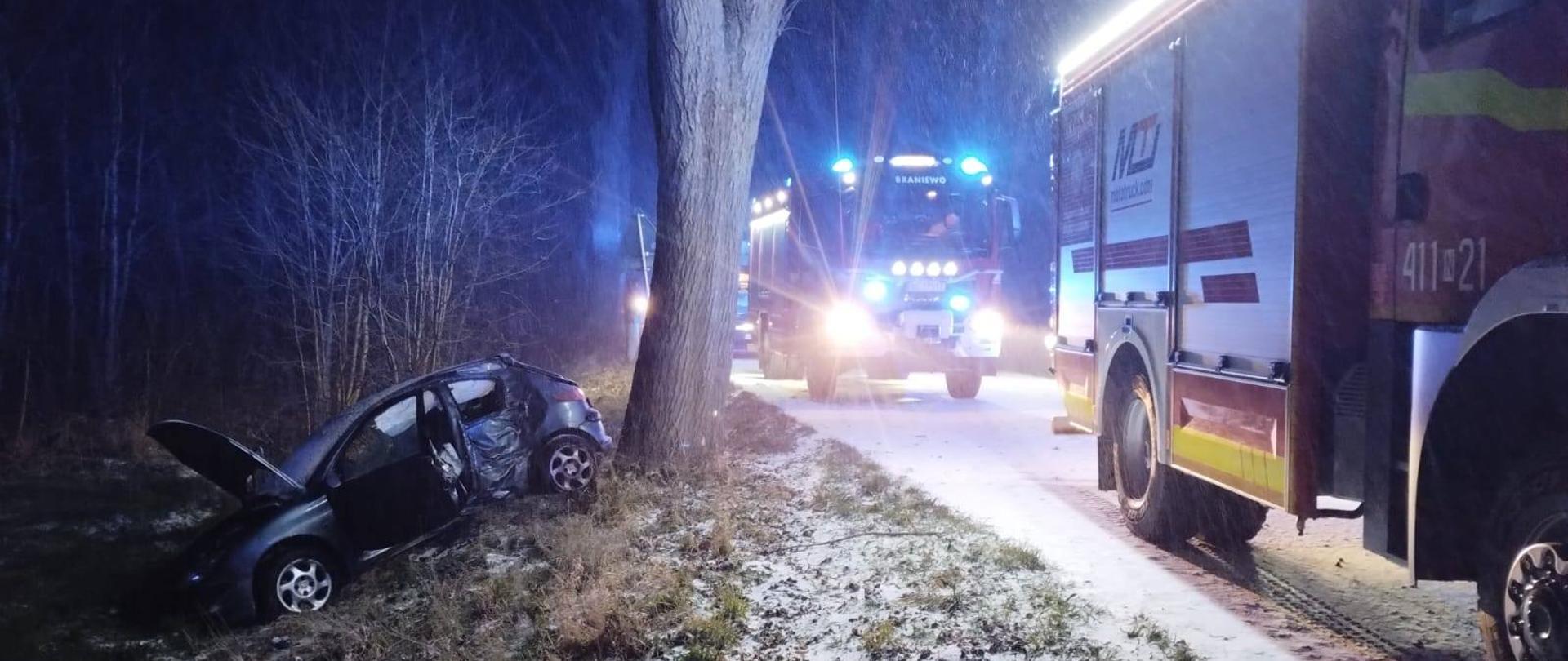 Zdjęcie zrobione nocą. Miejsce wypadku drogowego. Z lewej strony na poboczu drogi auto osobowe po uderzeniu w drzewo. Z prawej strony na drodze stoją dwa samochody straży pożarnej na światłach, ostawiając teren akcji i światłach ostrzegawczych.