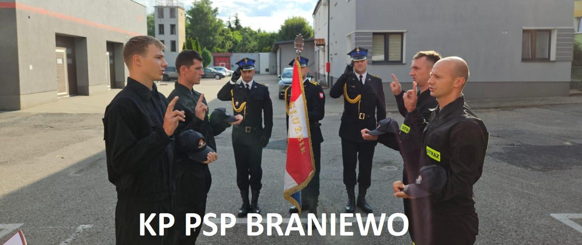Na zdjęciu strażacy. Po dwóch z prawej i lewej strony stoją do ślubowania z uniesiona dłonią. Na wprost poczet sztandarowy, strażacy w mundurach galowych. W tle budynki.