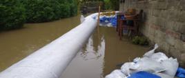 Na zdjęciu woda zalewa teren posesji oraz podchodzi pod budynek jednorodzinny. Wokół budynku ustawiono rękaw przeciwpowodziowy odgradzający wodę od budynku.