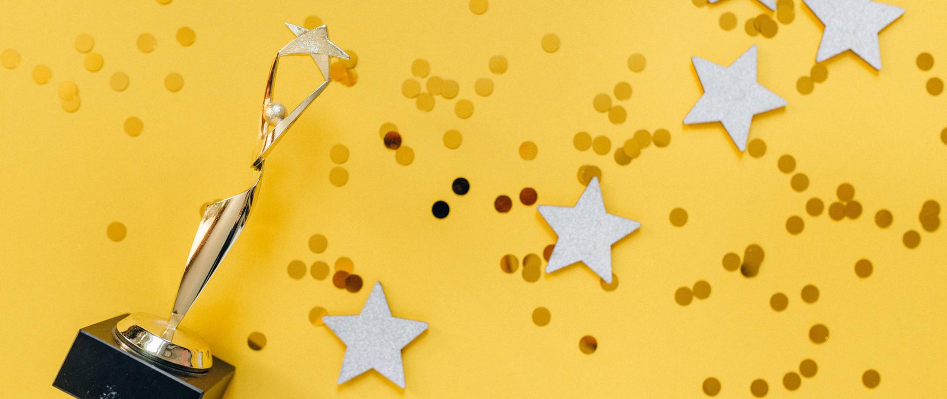 fotografia przedstawiająca złotą statuetkę postaci trzymającej gwiazdę, złote cekiny oraz pięć gwiazd rozrzuconych na żółtym tle
