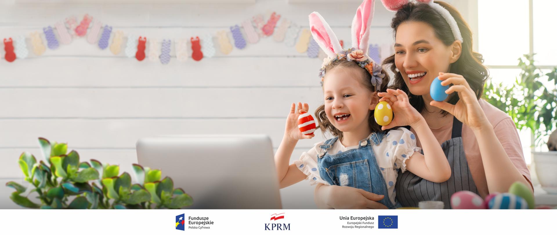 Uśmiechnięte mama i córka podczas wideorozmowy prze laptopa, pokazują kolorowe pisanki. Wokół nich wielkanocne ozdoby.