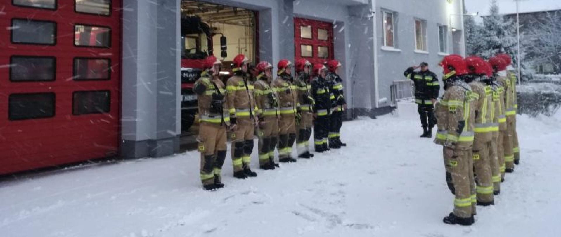 Zdjęcie przedstawia uroczystą zmianę służby. Zmiana zrobiona przed budynkiem komendy. Na zdjęciu naprzeciwko siebie stoją dwie zmiany służbowe. Jedna z bram otwarta i widać w niej pojazd gaśniczy. Tło zdjęcia ośnieżone, zmiana odbywa się podczas intensywnych opadów śniegu