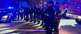 Policjanci i strażacy stoją przed swoimi samochodami służbowymi, część z nich salutuje. W tle lampy oświetlenia ulicznego, znaki drogowe. Na ziemi leży śnie, aura nocna.