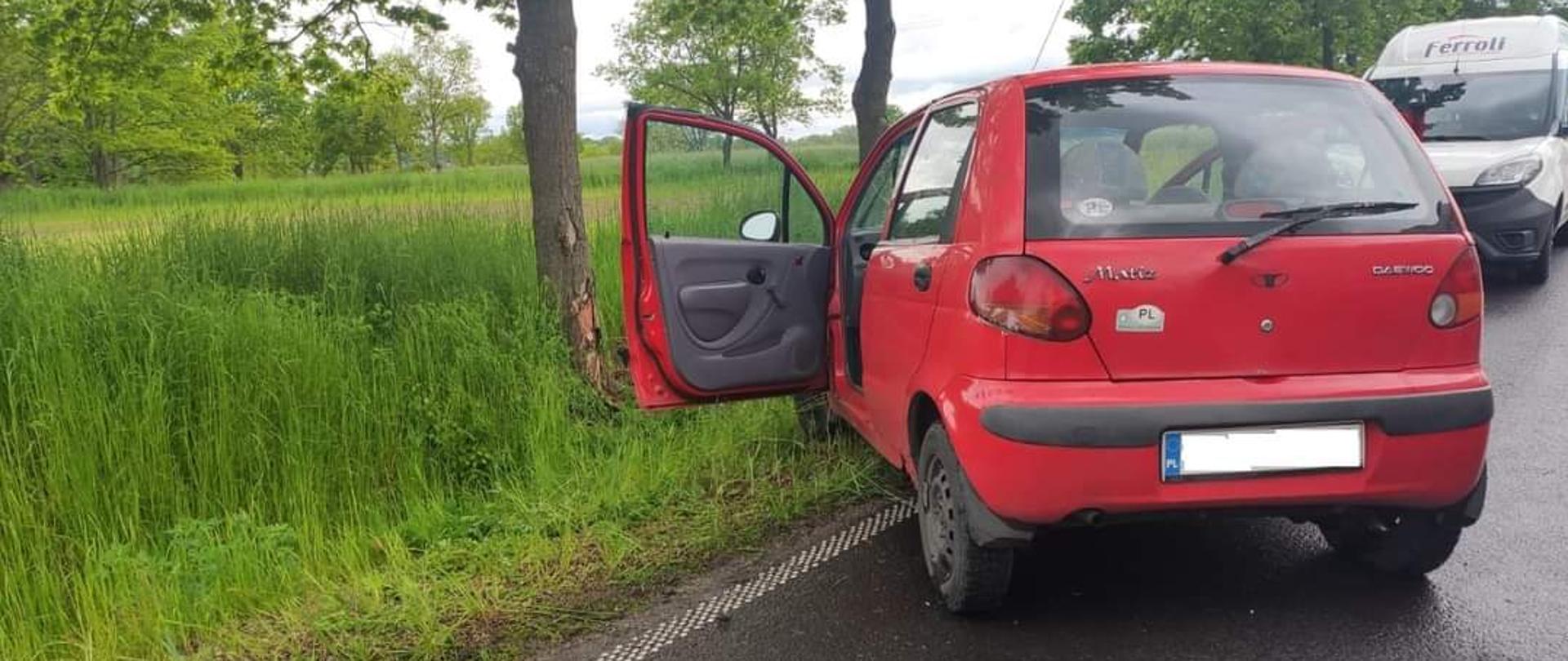 Pobocze drogi. Częściowo na pasie drogi stoi samochód osobowy Daewoo Matiz w kolorze czerwony. Drzwi kierowcy otwarte. W tle za przydrożnym rowem drzewo z częściowo zdartą korą. Za Matizem stoi biały bus.