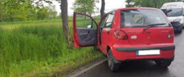 Pobocze drogi. Częściowo na pasie drogi stoi samochód osobowy Daewoo Matiz w kolorze czerwony. Drzwi kierowcy otwarte. W tle za przydrożnym rowem drzewo z częściowo zdartą korą. Za Matizem stoi biały bus.