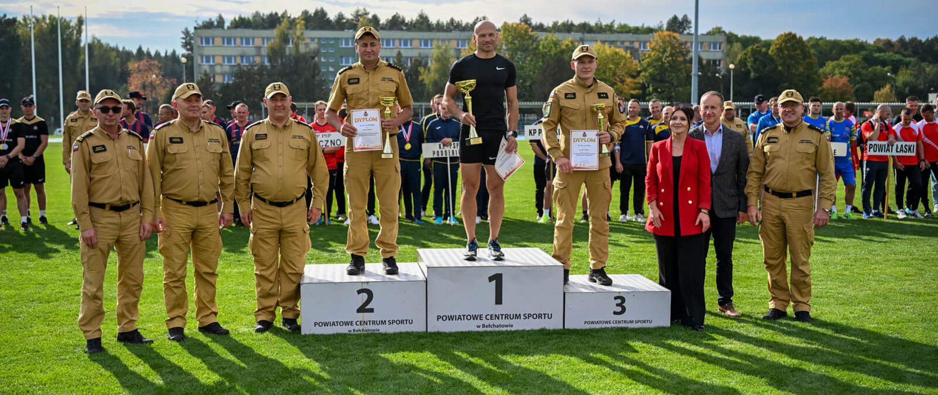 Laureaci klasyfikacji drużynowej stoją na podium z pucharami i dyplomami w otoczeniu strażaków w mundurach służbowych oraz zaproszonych gości.