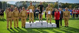 Dekoracja medalowa na podium. Na stopniach stoją trzej sportowcy w medalach, a po bokach asystują im strażak w mundurze oraz cywilni przedstawiciele.