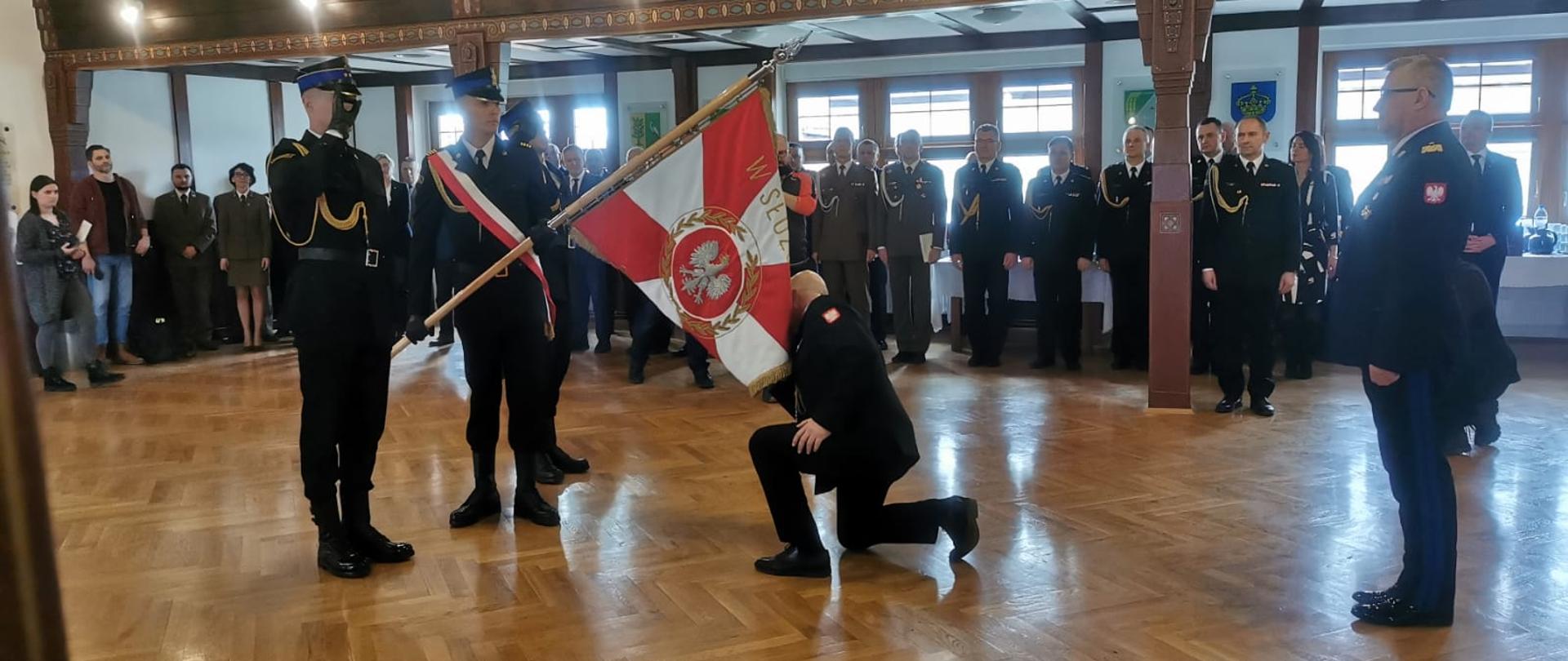 Na środku sali poczet sztandarowy pochyla sztandar. oficer PSP w mundurze galowym klęka, całuje płat sztandaru. Wokół zgromadzeni goście.