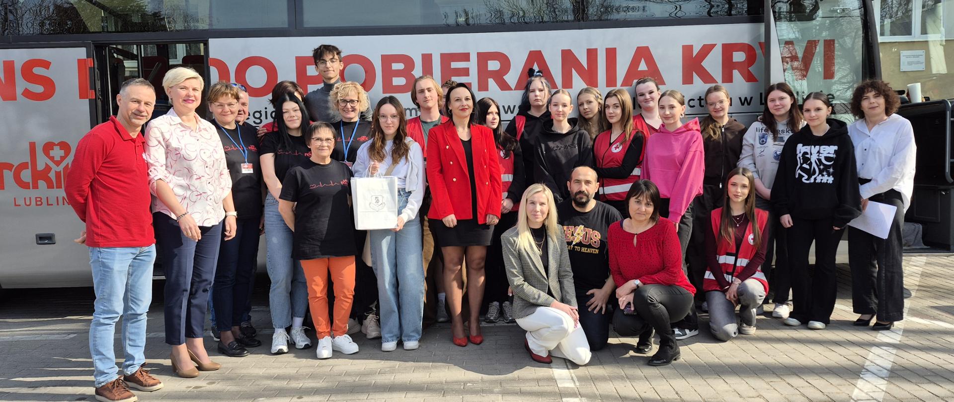 Grupa osób pozuje do wspólnego zdjęcia przed mobilnym punktem poboru krwi, stojąc na parkingu przed autobusem 