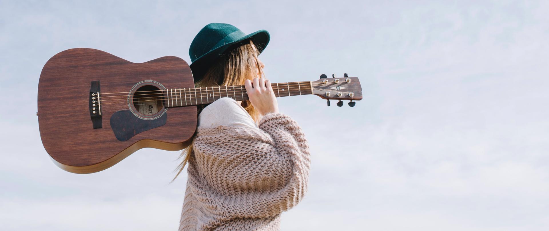 Fotografia przedstawia jasnowłosą kobietę z zielonym kapeluszem na głowie ubraną w kremowy sweter, która trzyma gitarę opartą na ramieniu, kobieta zwrócona jest w prawą stronę