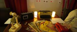 Na stoliku stoi magnetofon kasetowy Grundig, dwa telefony stacjonarne, maszyna do pisania oraz leżą gazety z lat 80-tych: Przekrój, Kobieta i Życie, Świat Młodych, Kontynenty i Szpilki