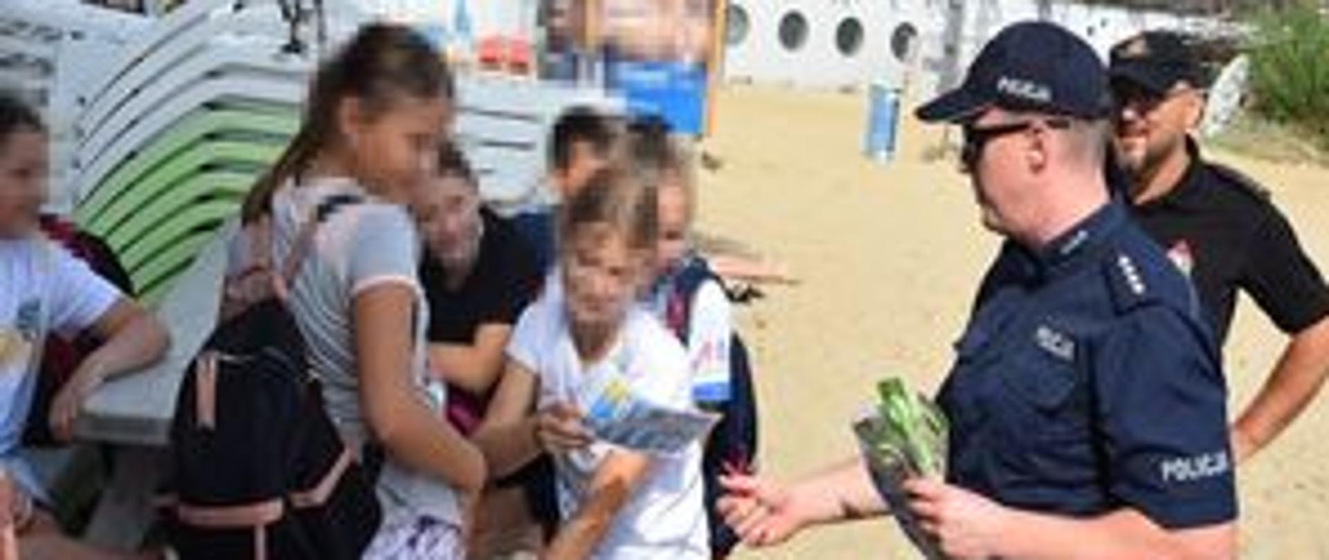 Na zdjęciu widać funkcjonariuszy Policji i Państwowej Straży Pożarnej wręczających ulotki i opaski dzieciom przebywającym na kolonii na puckiej plaży.