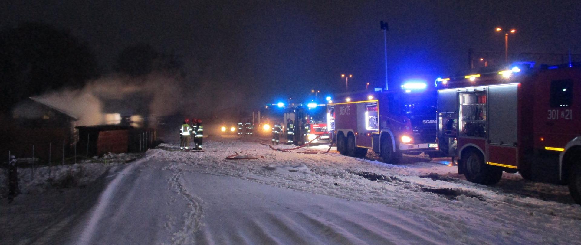 Droga pokryta śniegiem, trzech strażaków na środku zdjęcia, z prawej strony 4 wozy bojowe straży pożarnej z lewej zadymienie unoszące się z palącej się altany, nie widać płomieni.