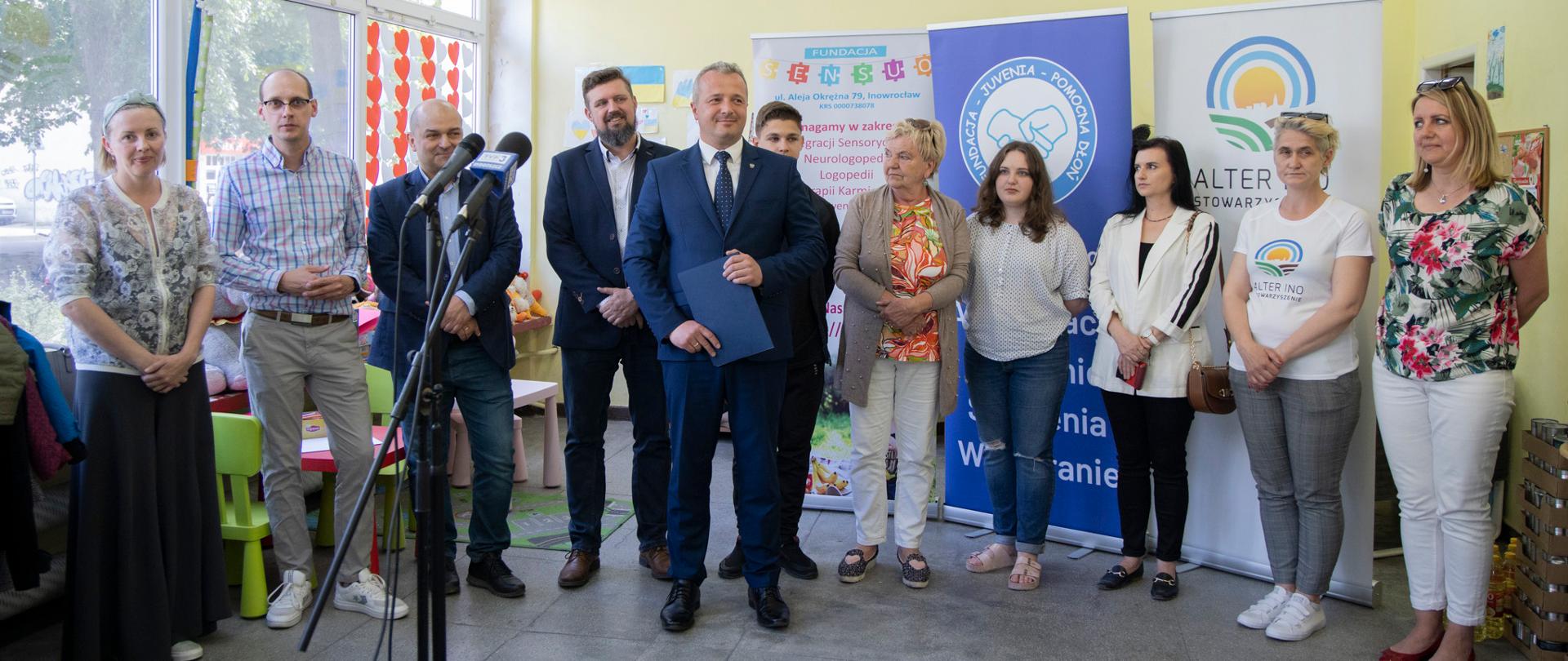 
Grupa ludzi stojąca w pomieszczeniu na tle roll-upów i plakatów
