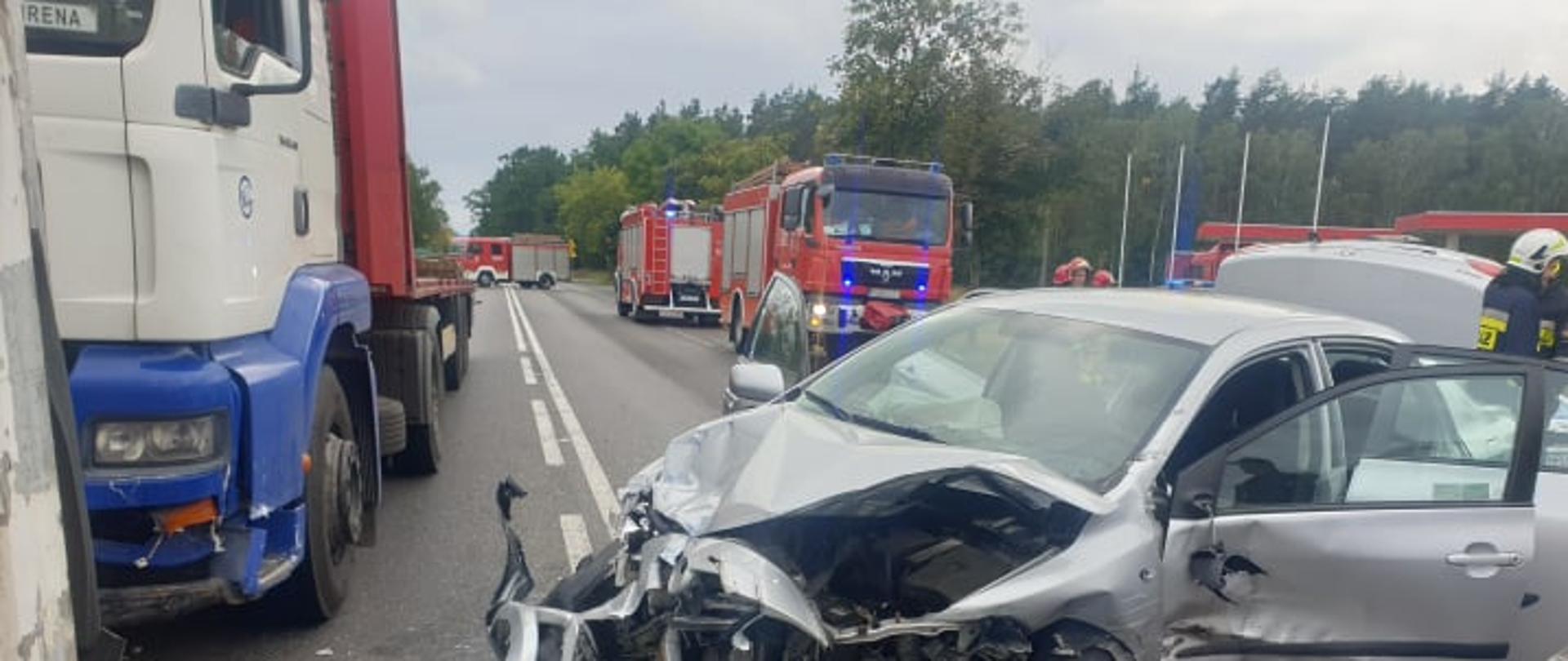 Zdjęcie przedstawia rozbity samochód osobowy marki Toyota Corolla na tle pojazdów strażackich oraz tira.