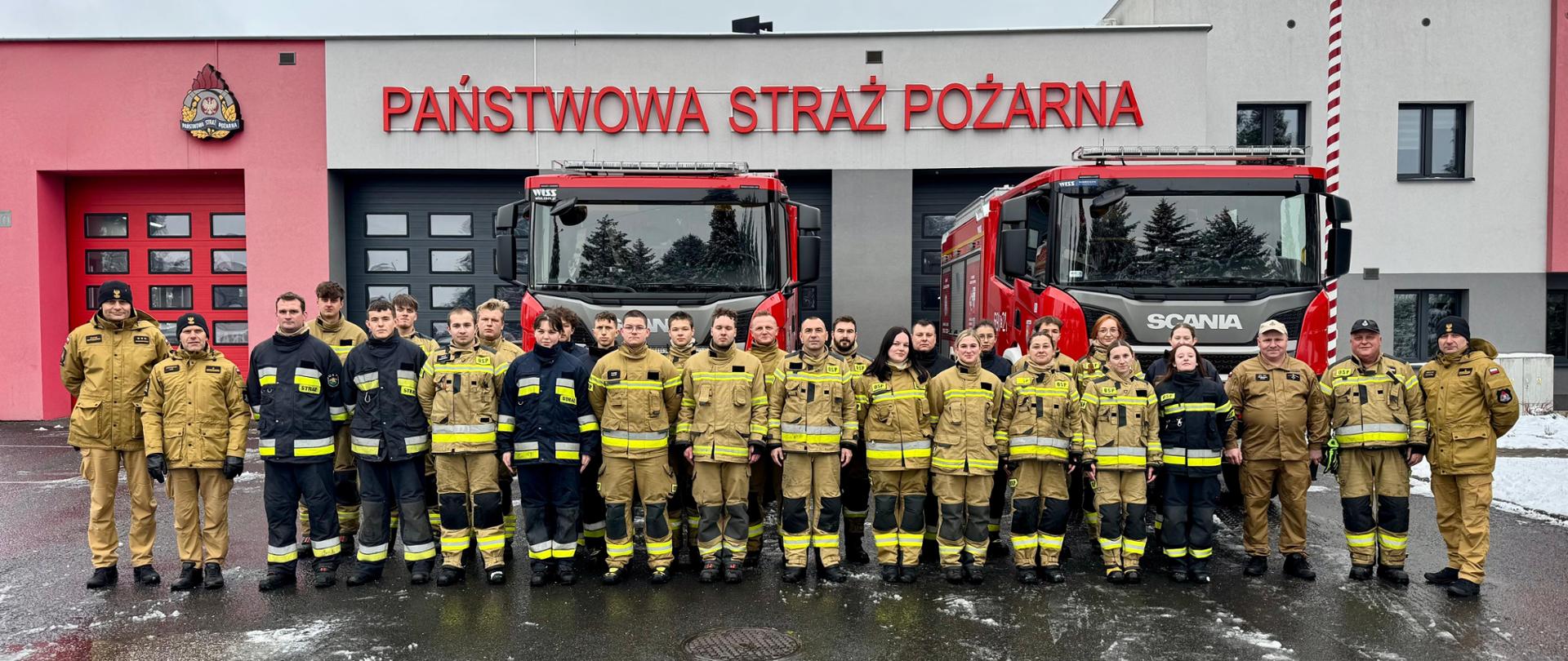 Grupa strażaków w pełnym umundurowaniu, stojących w szeregu przed budynkiem remizy strażackiej. Na fasadzie budynku widnieje napis „PAŃSTWOWA STRAŻ POŻARNA”. Przed remizą zaparkowane są dwa czerwone wozy strażackie marki Scania. Strażacy mają na sobie hełmy, kurtki ochronne i spodnie, a ich postawa jest wyprostowana, skierowana w stronę aparatu. Na ziemi widać wilgoć, a po bokach zalega śnieg.