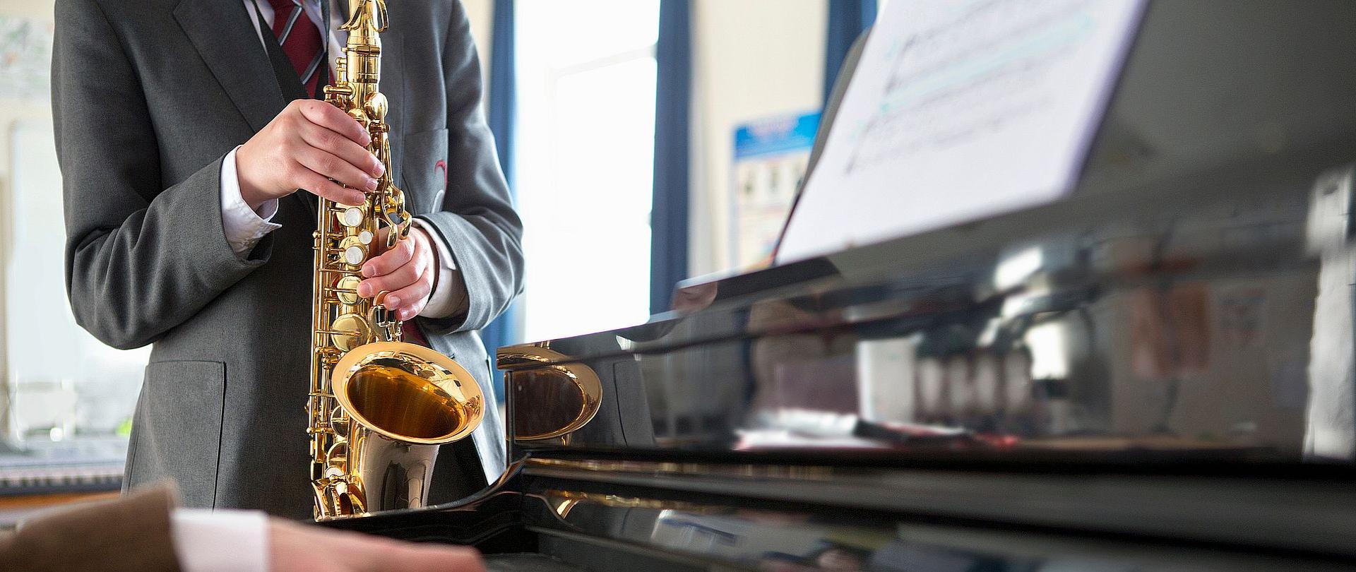 Zdjęcie przedstawia osobę ubraną w garnitur i grającą na saksofonie. Na pierwszym planie zdjęcia znajdują się ręce akompaniującego pianisty.