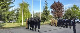 Strażacy stoją w dwóch szeregach przy maszcie flagowym. Przy maszcie stoi poczet flagowy. Po środku stoi strażak w mundurze wyjściowym i salutuje. W tle drzewa i samochody.
