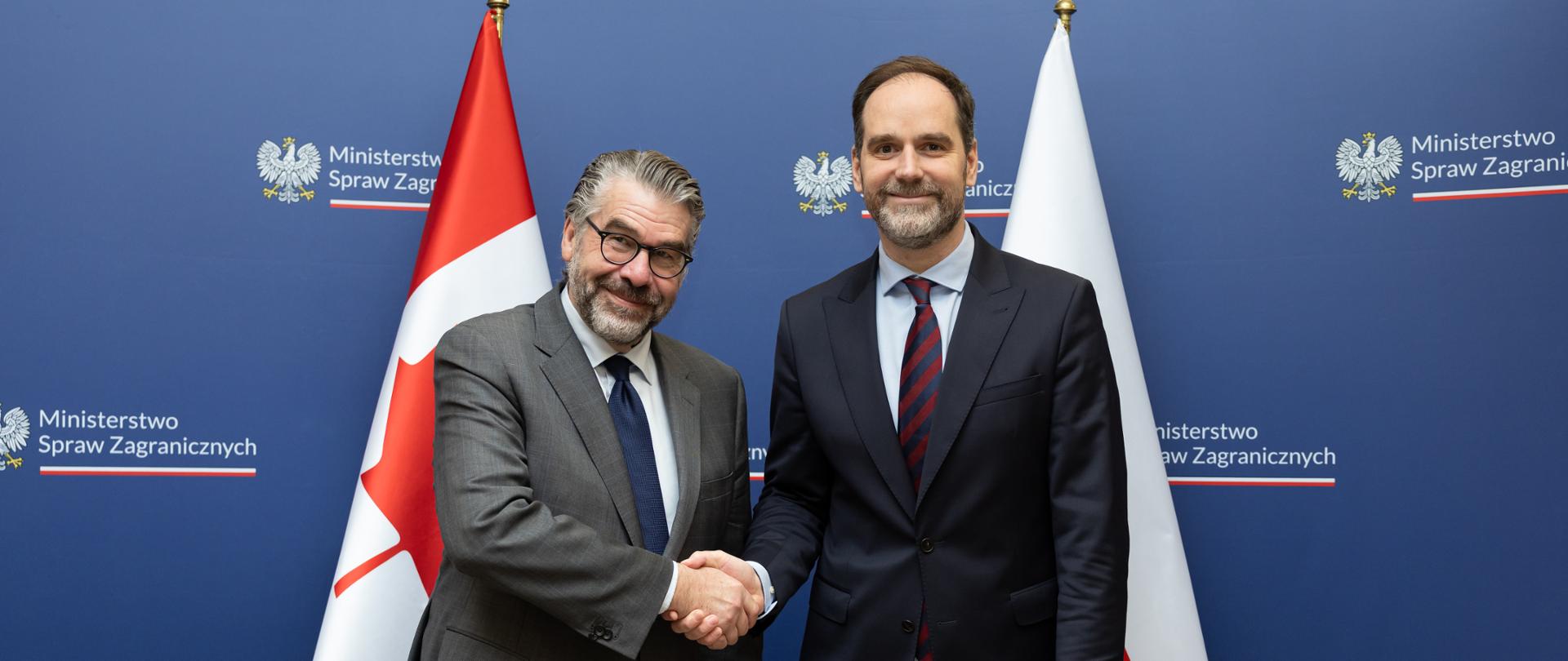 Na zdjęciu jest dwóch mężczyzn w garniturach. Za nimi jest ścianka z logo MSZ i dwie flagi - Kanady i Polski. 